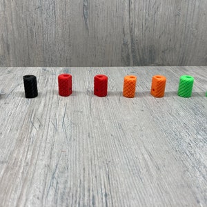 May include: Eight different colored cylindrical knobs with a textured surface. The knobs are arranged in a row from left to right, starting with black, then red, orange, and green.