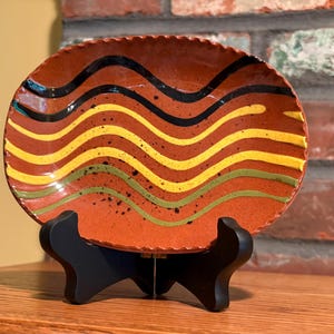 May include: An oval ceramic dish with a scalloped edge, featuring a terracotta-colored base decorated with horizontal wavy lines in black, yellow, and olive green, accented by small black speckles, displayed on a black plastic tripod stand.
