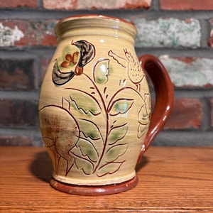 May include: A cream-colored ceramic mug with a glossy finish, featuring a hand-painted design of a bird, a flower, and a deer in shades of green, brown, and black. The mug has a dark reddish-brown handle and a matching base rim.