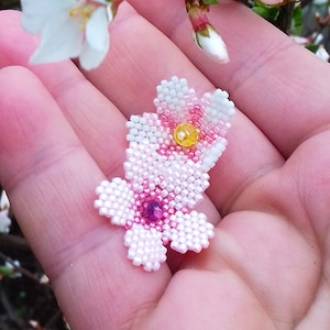May include: A close-up of a hand holding a beaded pink and white flower. The flower is a beaded replica of a cherry blossom. The text "Sakura flowers 23T002" is below the image.