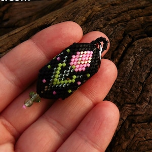 May include: A close-up of a small, pink and green beaded tulip flower pendant. The pendant is made using the peyote stitch and is held in a hand against a wooden background. The text "Tulips 20T001" is below the image.
