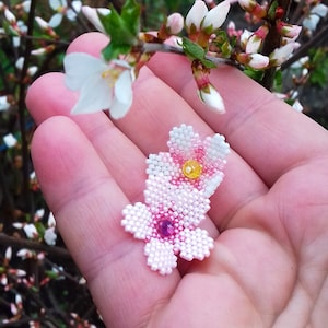 May include: A close-up of a pink and white beaded flower, resembling a cherry blossom, held in a hand against a background of green leaves. The flower is a peyote stitch beading project, with a yellow center bead. The image includes the text "Sakura flowers 23T002" and "Peyote tutorial for beading pattern in PDF | Original design 2023 &copy; Lady Lunar Cat".