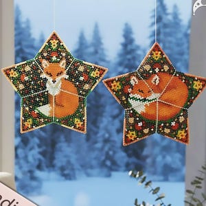 May include: Two star-shaped beaded ornaments with fox designs. The foxes are orange with white accents, set against a dark background with floral and geometric patterns. The ornaments are suspended in front of a snowy winter scene. The text "Beading pattern" is visible.