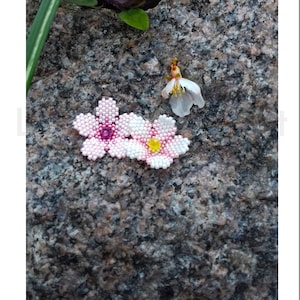 May include: Two beaded cherry blossom flowers, one pink with a pink center bead and one white with a yellow center bead, on a gray and brown textured rock surface. The text "Sakura flowers 23T002" is below the image.