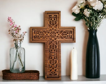 May include: A large, intricately carved wooden cross with a detailed floral design. The cross is mounted on a white wall and is surrounded by a vase of white flowers and a white candle.
