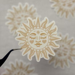 May include: A beige sun sticker with a smiling face, closed eyes, and heart-shaped cheeks. The sun has radiating rays and is held by tweezers. Other similar stickers are in the background.