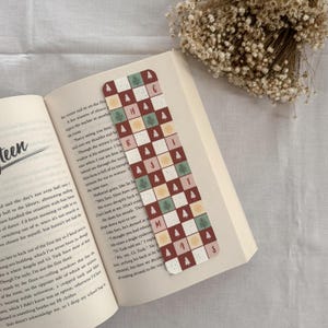 May include: A decorative bookmark with a Christmas theme. The bookmark features a checkered pattern with red, white, and green squares, each adorned with a Christmas tree or a letter spelling "CHRISTMAS". It is placed in an open book.