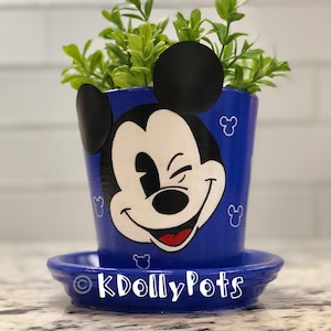 May include: Blue ceramic planter with a painted image of Mickey Mouse winking. The planter has a saucer and is filled with green plants. The text "KDolleyPots" is on the saucer.