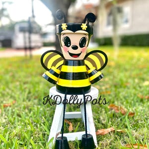May include: A whimsical, hand-painted bee-themed flower pot. The pot features a smiling face, black and yellow stripes, and decorative wings. The pot is sitting on a small white stool. The text "KDolly Pots" is visible on the stool.