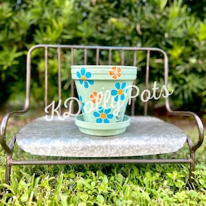 May include: A light green terracotta flower pot with a blue and orange floral design. The pot is sitting on a metal bench with a galvanized finish.