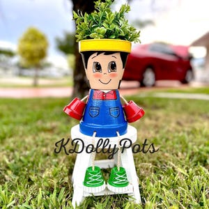 May include: A hand-painted terracotta flower pot shaped like a boy, with a yellow brim, red hands, and blue overalls. The pot is filled with green plants and sits on a white stool. The text "KDolly Pots" is visible.