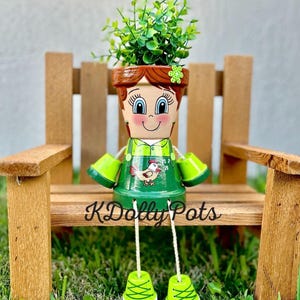 May include: A terracotta pot figure with a painted face, brown hair, and a green outfit featuring a chicken graphic. The figure has dangling legs with green shoes, arms made of small pots, and holds a green plant. The text KDolly Pots is visible.