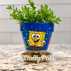 May include: A blue and yellow hand-painted terracotta flower pot with a cartoon image of Spongebob Squarepants. The pot has a white saucer and is filled with green leafy plants.