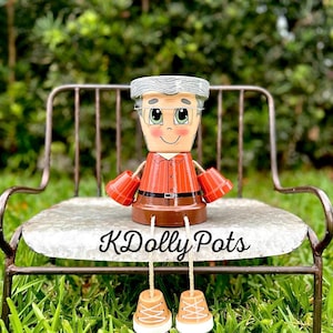 May include: A decorative terracotta pot figure featuring a painted face with gray hair and glasses, wearing an orange collared shirt and brown pants. The figure has small terracotta pot hands and feet, with the text KDolly Pots printed on the seat.