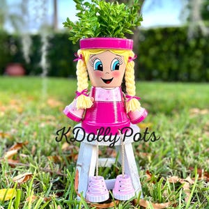 May include: A hand-painted flower pot shaped like a girl with blonde braids, wearing a pink dress and shoes, and holding a plant. The pot is sitting on a small white stool. The text "K Dolly Pots" is visible.