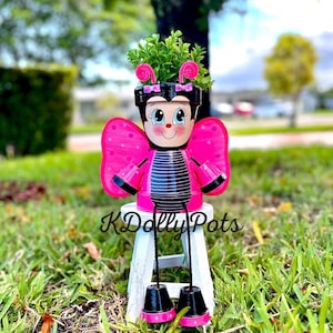 May include: A decorative planter crafted from stacked terracotta pots painted to resemble a butterfly character with a black and pink striped body, large pink wings, and a smiling face. The planter features pink pipe cleaner antennae and holds a small green plant, resting on a white wooden stool with the text KDolly Pots.