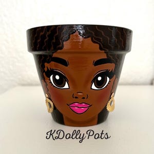 May include: A terracotta plant pot hand-painted with the face of a person with dark skin, wavy dark hair, and bright pink lips. The character wears gold-colored fan-shaped earrings. The text KDolly Pots is printed in black below the pot.