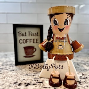 May include: A decorative figurine crafted from terracotta pots, featuring a painted face with a brown braided ponytail, wearing a tan apron with the text But FIRST Coffee and a coffee cup graphic. The figure has dangling legs with brown shoes.