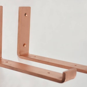 May include: Two copper shelf brackets with a 90-degree angle and a curved end. The brackets are made of metal and have a brushed finish.