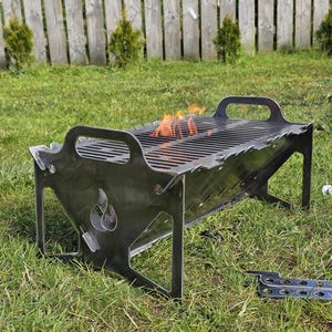 May include: A portable, rectangular metal fire pit and grill featuring a dark grey finish, integrated side handles, and a flame cutout design on the end panels. The unit has a slatted cooking grate with visible orange flames burning inside.