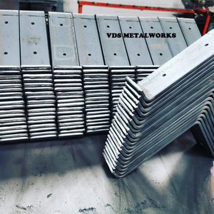 Op de afbeelding: Een stapel metalen beugels, sommige recht en sommige gebogen in een hoek van 90 graden. De beugels zijn allemaal zilverkleurig en hebben meerdere gaten die erin zijn geboord. De tekst "VDS METALWORKS" is zichtbaar op de bovenkant van de stapel.