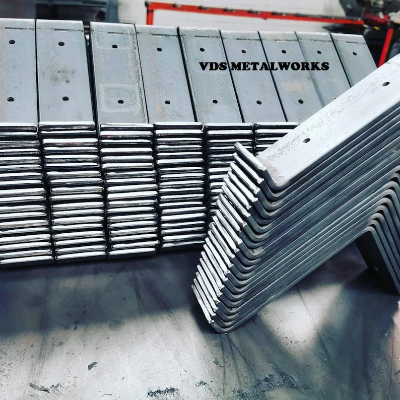 Op de afbeelding: Een stapel metalen beugels, sommige recht en sommige gebogen in een hoek van 90 graden. De beugels zijn allemaal zilverkleurig en hebben meerdere gaten die erin zijn geboord. De tekst "VDS METALWORKS" is zichtbaar op de bovenkant van de stapel.