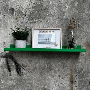 May include: A green metal shelf with a white picture frame, a small potted plant, and a glass jar with a plant inside. The picture frame has the word "HAPPY" spelled out with wooden letter tiles. The text below the picture reads "There are so many beautiful reasons to be happy."