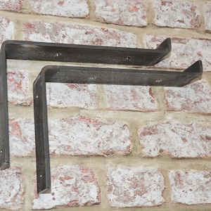 Op de afbeelding: Twee zwarte metalen plankbeugels gemonteerd op een bakstenen muur. De beugels zijn L-vormig en hebben een rustieke afwerking.