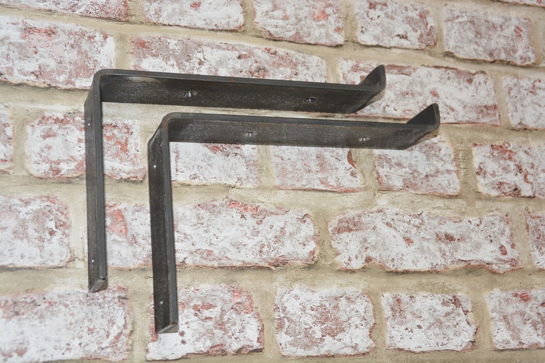 Op de afbeelding: Twee zwarte metalen plankbeugels gemonteerd op een bakstenen muur. De beugels zijn L-vormig en hebben een rustieke afwerking.