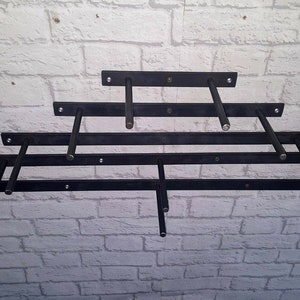 Puede incluir: Cinco soportes de estantería de metal negro montados en una pared de ladrillo blanco. Los soportes están dispuestos en forma escalonada, con el soporte más largo en la parte inferior y el más corto en la parte superior.