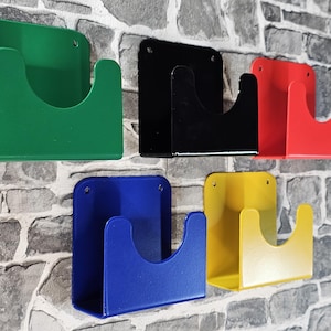 May include: Six wall-mounted metal holders in green, black, red, blue, and yellow. Each holder has a rectangular backplate and a curved front section. The holders are mounted on a grey and white brick wall.