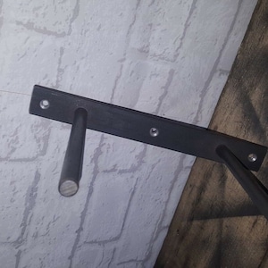 May include: Black metal shelf brackets with two arms extending out from a rectangular base. The brackets are mounted to a white brick wall and a wooden surface.