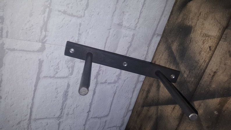 May include: Black metal shelf brackets with two arms extending out from a rectangular base. The brackets are mounted to a white brick wall and a wooden surface.