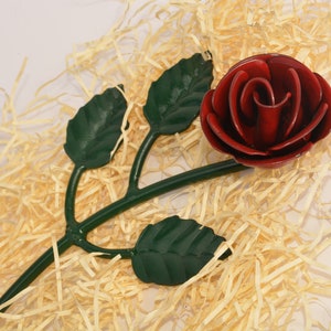 May include: A handcrafted metal rose with a deep red bloom and green stem and leaves. The rose is set against a bed of light-colored straw, creating a rustic and elegant display. A decorative floral accent.