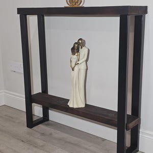 May include: A dark brown wooden console table with black metal legs. A gold candle holder and a white figurine of a couple are displayed on the table. The table is against a white wall and sits on a light grey floor.