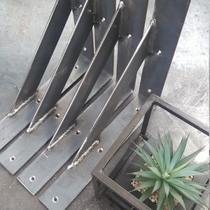 Könnte beinhalten: Satz von vier dunklen Metall-Winkelhaltern mit abgewinkelten Stützen, jeweils mit vorgebohrten Löchern zur Montage. Ein kleiner, quadratischer Metall-Pflanzkasten mit einer künstlichen Sukkulentenpflanze ist ebenfalls vorhanden.