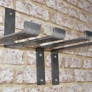 Op de afbeelding: Vier metalen plankdragers gemonteerd op een bakstenen muur. De dragers zijn gemaakt van stevig metaal en hebben een robuuste, industri&euml;le uitstraling.