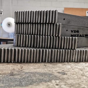 Op de afbeelding: Een stapel metalen tandwielen met een VDS Metalworks-logo op het bovenste stuk. De tandwielen zijn gemaakt van donkergrijs metaal en hebben een ruwe, industri&euml;le uitstraling.