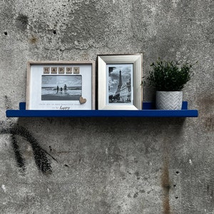 Puede incluir: Dos fotos enmarcadas en un estante azul contra una pared de concreto gris. La foto de la izquierda tiene la palabra "HAPPY" deletreada en fichas de madera. La foto de la derecha muestra una foto en blanco y negro de la Torre Eiffel. Una pequeña planta en maceta se encuentra en el estante entre las fotos.