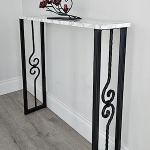 May include: A console table with a marble effect top and black metal legs. The legs have decorative scrollwork. A metal sculpture of red flowers and a heart sits on the table. The table is against a white wall.