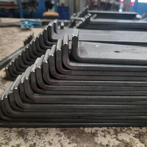 Op de afbeelding: Een stapel L-vormige metalen beugels, waarschijnlijk gebruikt voor bouw- of doe-het-zelfprojecten. De beugels zijn gemaakt van donkergrijs metaal en hebben een glad, licht getextureerd oppervlak.
