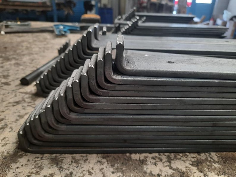 Op de afbeelding: Een stapel L-vormige metalen beugels, waarschijnlijk gebruikt voor bouw- of doe-het-zelfprojecten. De beugels zijn gemaakt van donkergrijs metaal en hebben een glad, licht getextureerd oppervlak.