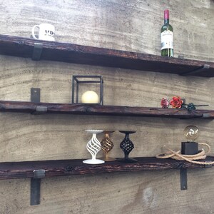 Op de afbeelding: Drie rustieke houten planken met metalen beugels, gemonteerd op een getextureerde muur. De bovenste plank heeft een witte mok en een fles wijn. De middelste plank heeft een zwarte metalen kubus met een witte kaars erin. De onderste plank heeft drie decoratieve kandelaars en een kleine gloeilamp op een snoer.