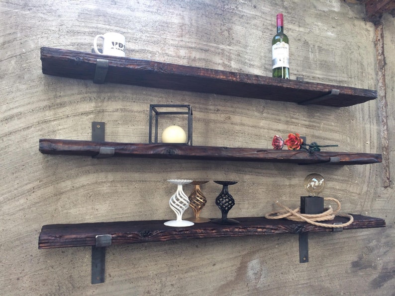 Op de afbeelding: Drie rustieke houten planken met metalen beugels, gemonteerd op een getextureerde muur. De bovenste plank heeft een witte mok en een fles wijn. De middelste plank heeft een zwarte metalen kubus met een witte kaars erin. De onderste plank heeft drie decoratieve kandelaars en een kleine gloeilamp op een snoer.
