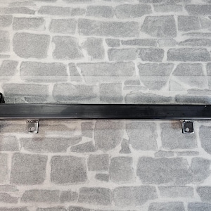 May include: A black metal shelf bracket for wall mounting. It has a rectangular bar supported by a mounting plate and two smaller brackets. The background is a grey brick wall.