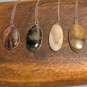 May include: Six gemstone pendant necklaces with silver chains. The pendants are oval and feature various colors and patterns, including brown, green, purple, and white. The necklaces are displayed on a wooden tray.