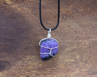 May include: A purple and black stone pendant wrapped in silver-colored wire, suspended from a black cord necklace. The stone has a textured appearance. The background is a wooden surface.