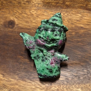 May include: A green and pink carved stone figurine of a whimsical character wearing a hat. The figurine has a smiling face and is holding a stick in its hand.