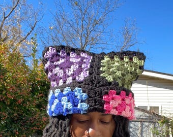 May include: A colorful crochet hat featuring a patchwork design. The hat incorporates squares of black, purple, blue, pink, and green yarn. The hat is handmade and appears to be a beanie style.