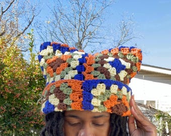 May include: A colorful crochet hat with a cat ear design. The hat features a patchwork pattern in shades of blue, orange, brown, and green. The hat is handmade and appears to be worn by a person.